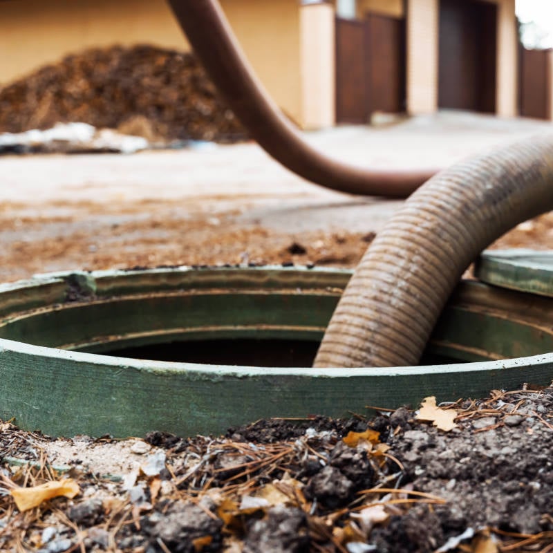 A large hose is inserted into an open outdoor septic tank, likely for septic tank pumping or maintenance, with dirt and leaves scattered around the area.