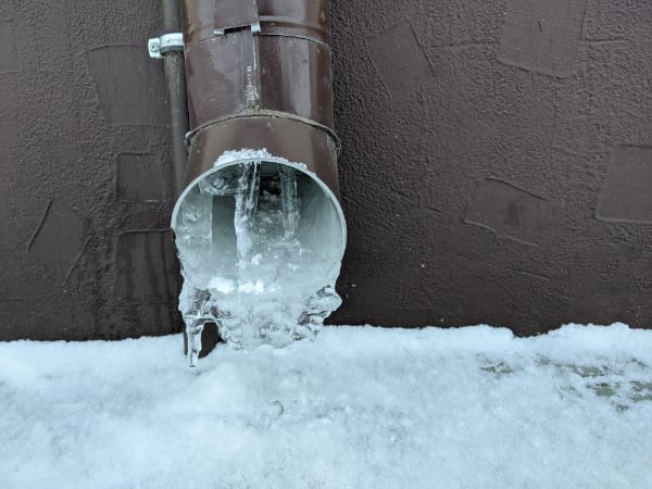 A brown metal drainpipe with icicles and ice buildup at the opening, attached to a textured wall above snow-covered ground—a clear reminder of the importance of preventing frozen pipes in winter.