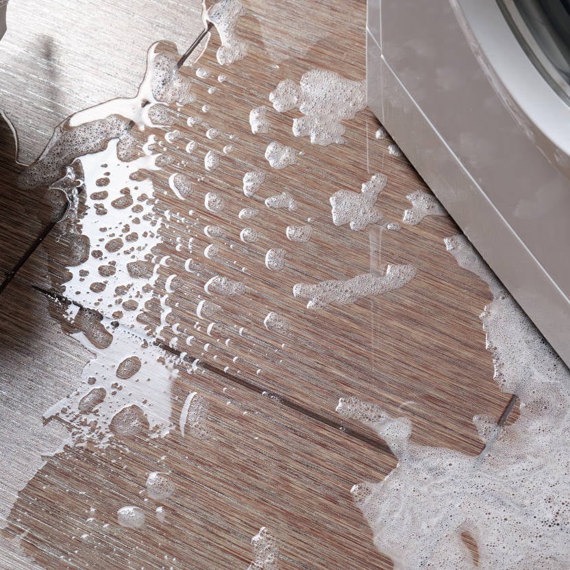 Soapy water spilling onto a wooden floor from the side of a washing machine, creating bubbles and puddles—a clear sign that drain cleaning may be needed.