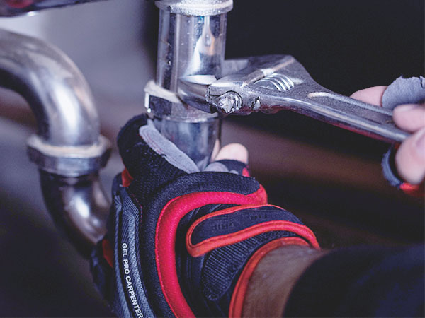 Person wearing gloves uses a wrench to tighten or loosen a metal pipe under a sink.