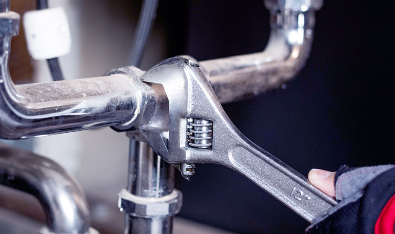 A hand uses a wrench to tighten or loosen a metal pipe fitting under a sink.