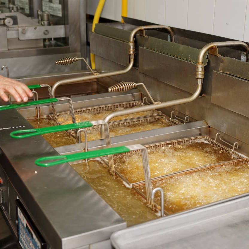 Commercial deep fryer with metal baskets submerged in hot oil, food frying inside. A hand holds one basket handle. Stainless steel surfaces and fryer controls are shown—essential equipment for kitchens that rely on regular Grease Trap Cleaning.