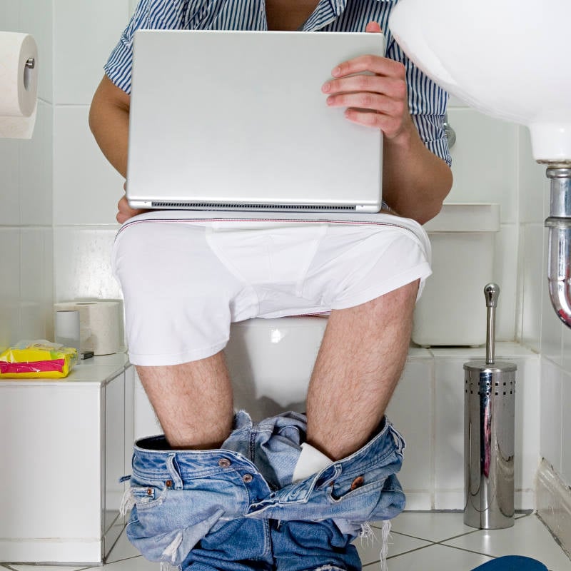 A person sits on a toilet with pants and underwear down, using a laptop in the bathroom—perhaps searching plumbing services to fix clogged toilets or handle urgent toilet repair needs.