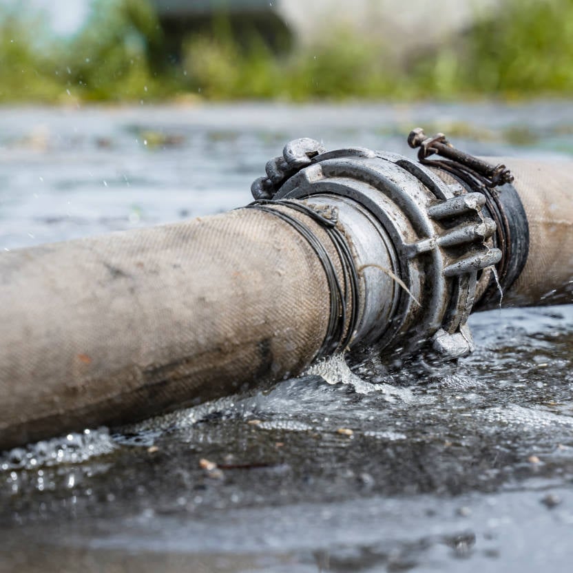 A large industrial hose is jetting water from a leak at the metal connector, causing water to pool on the ground.