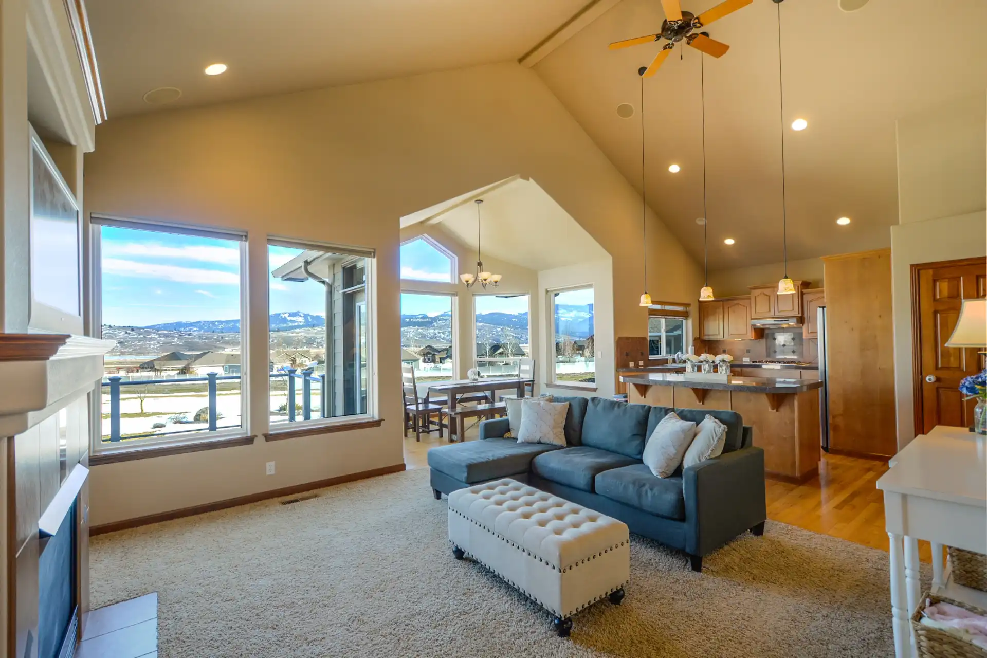 Open-concept living room and kitchen with vaulted ceilings, large windows showing a mountain view, modern furnishings, and a dining area near the windows.